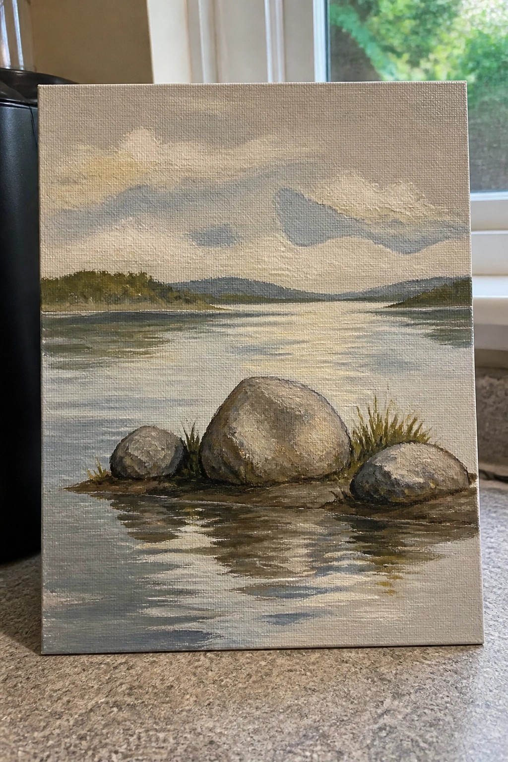 Realistic landscape painting of rocks in calm water reflecting cloudy hills and sky.