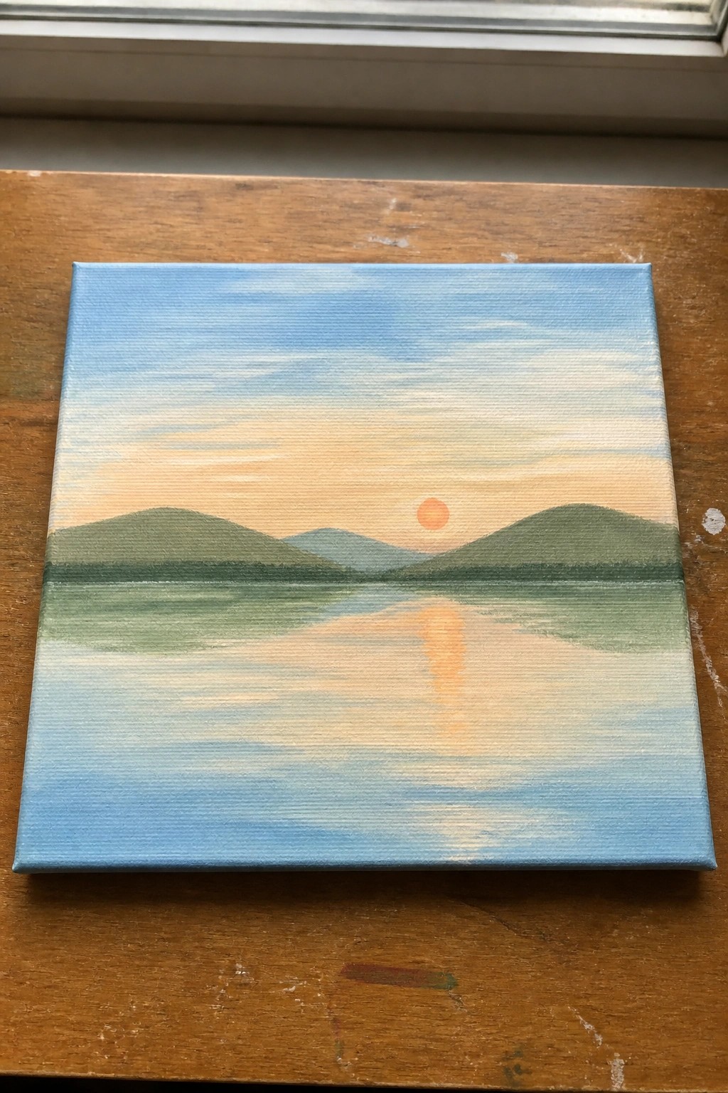 Canvas painting of sunset over green hills with reflection in calm water on wooden table