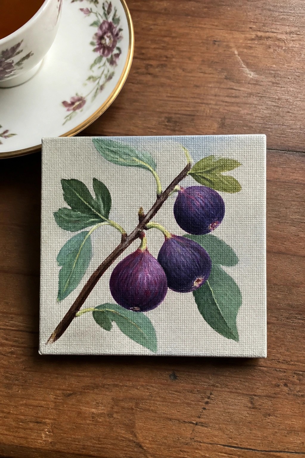 Small canvas painting of purple figs on branch beside floral teacup on wood.