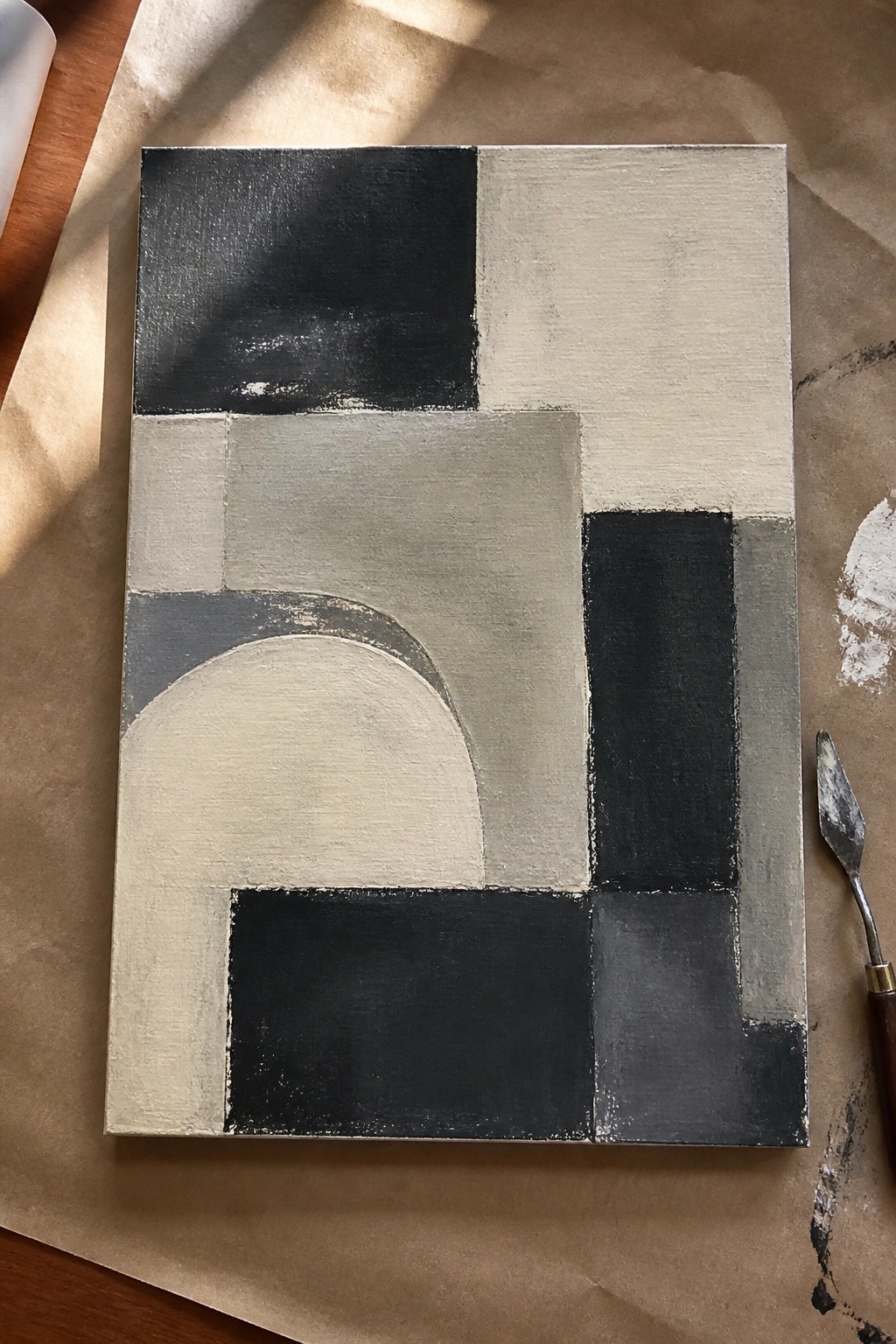 Abstract geometric painting in black, gray, and beige tones beside a palette knife on paper.