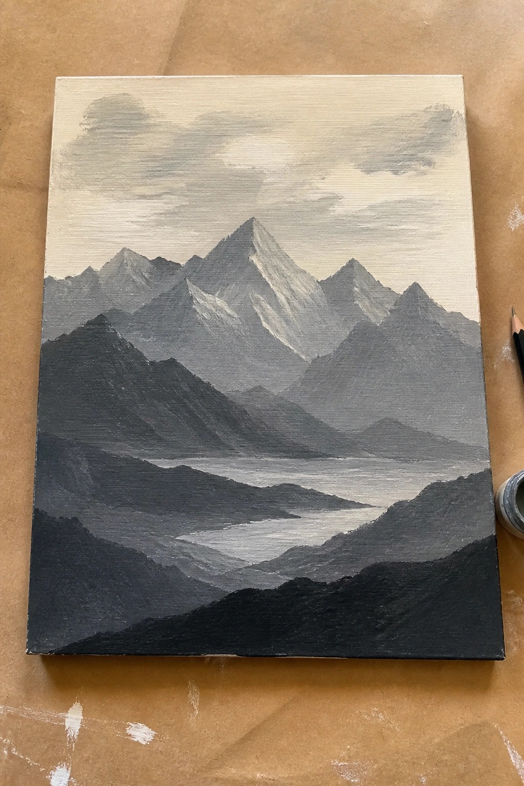 Grayscale mountain landscape painting with misty valley on canvas beside pencil