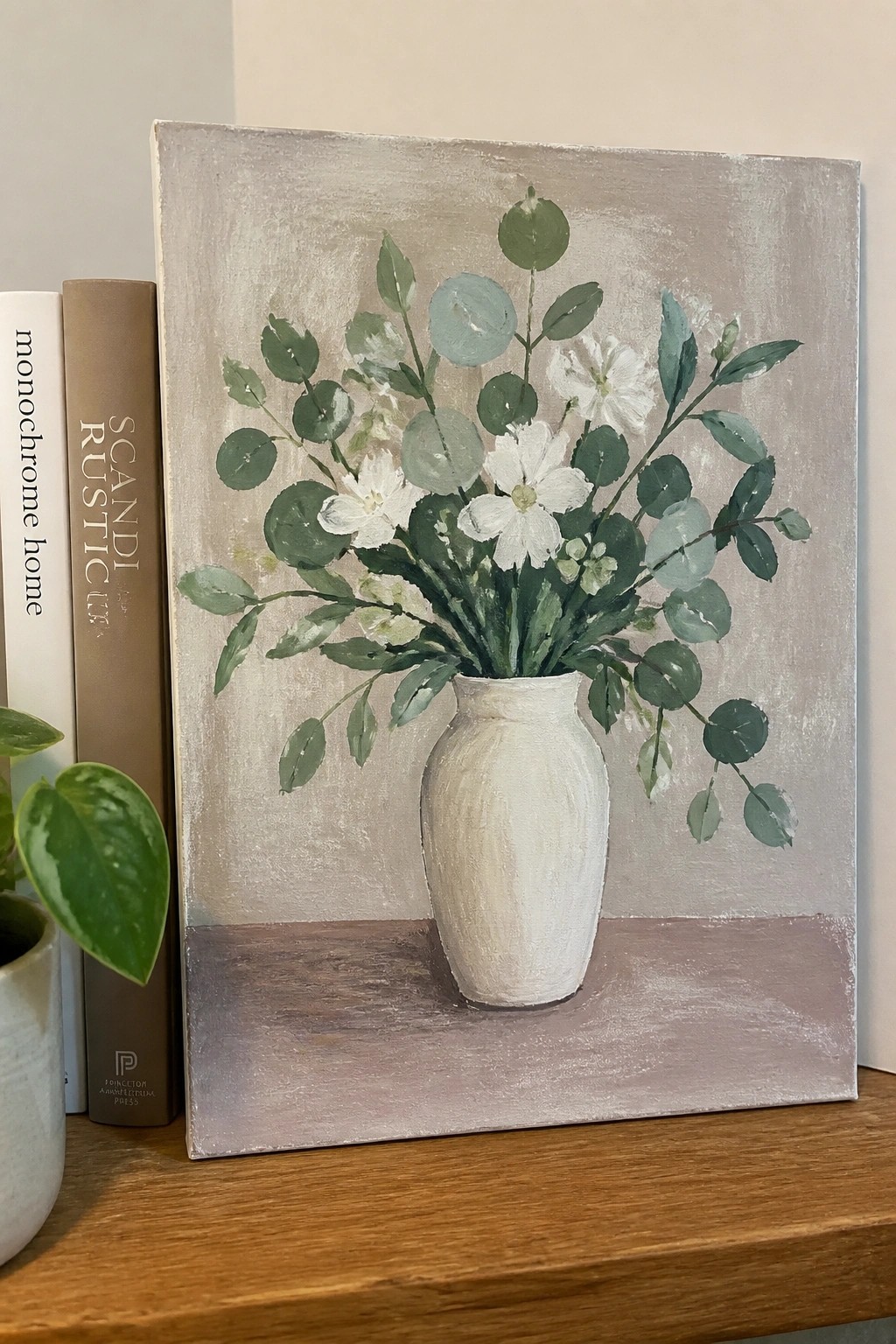 Still life painting of white flowers and green leaves in a white vase beside books.