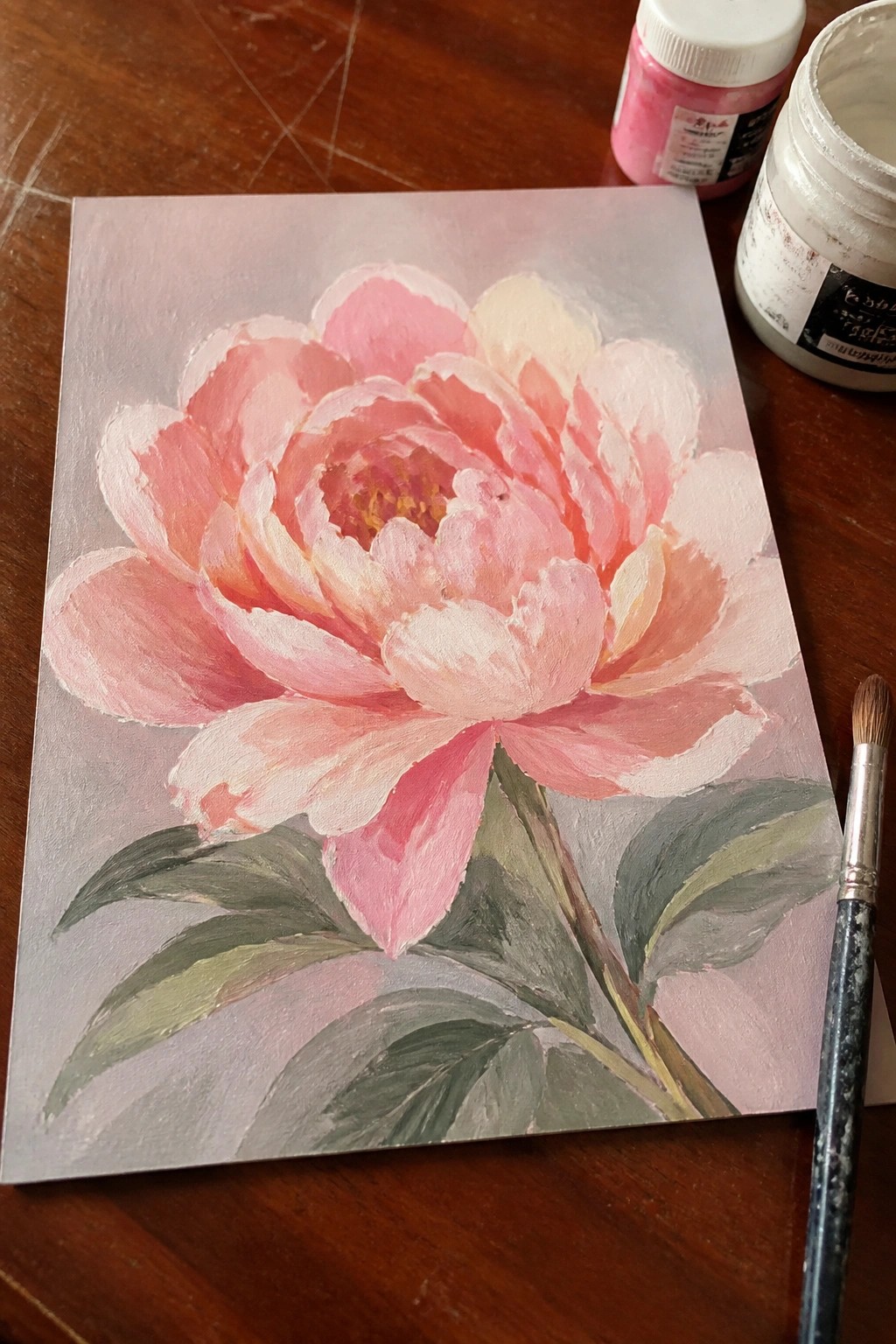 Pink peony painting on canvas beside paint jars and brush on wooden table