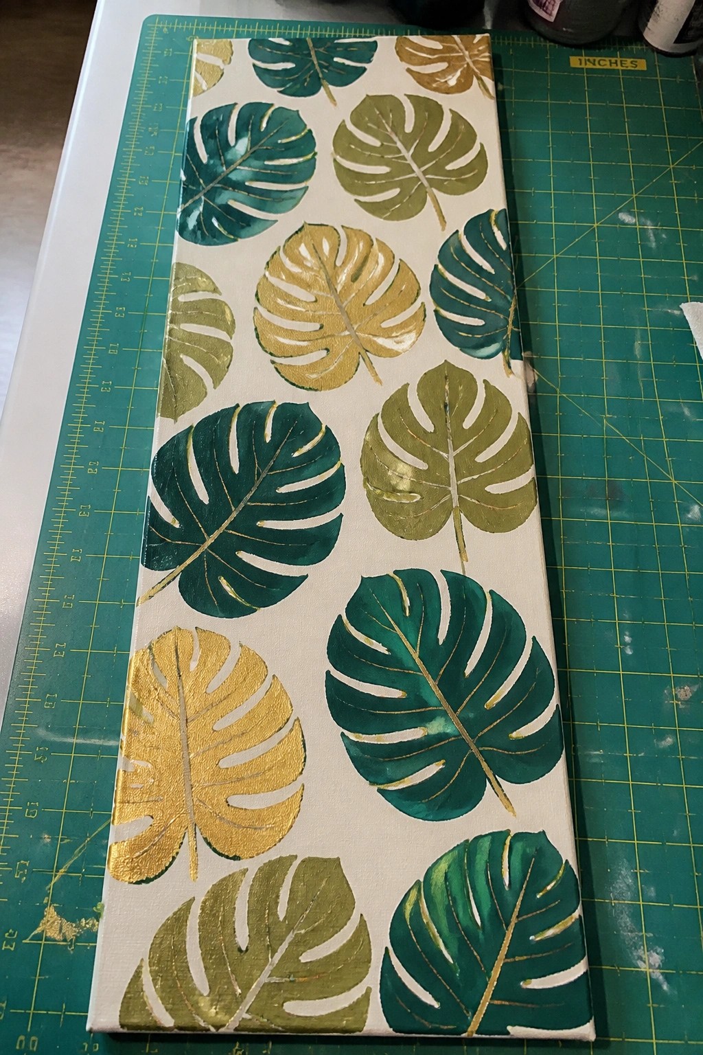Canvas with green and gold monstera leaves on green cutting mat.