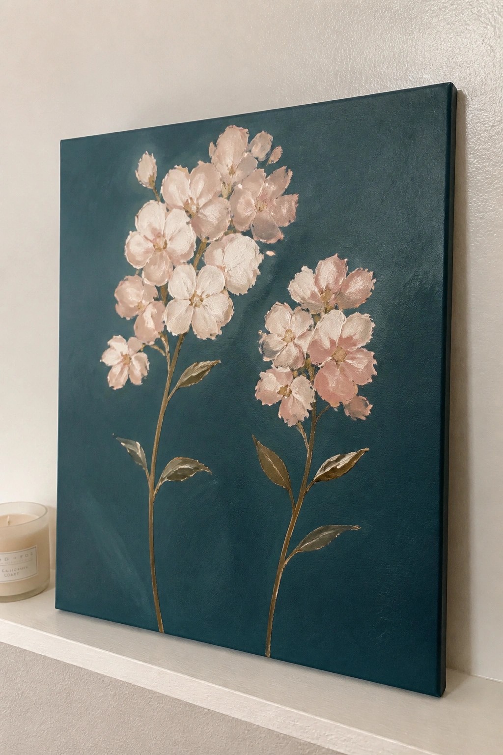 Painting of pink flowers with gold stems on dark teal canvas beside candle