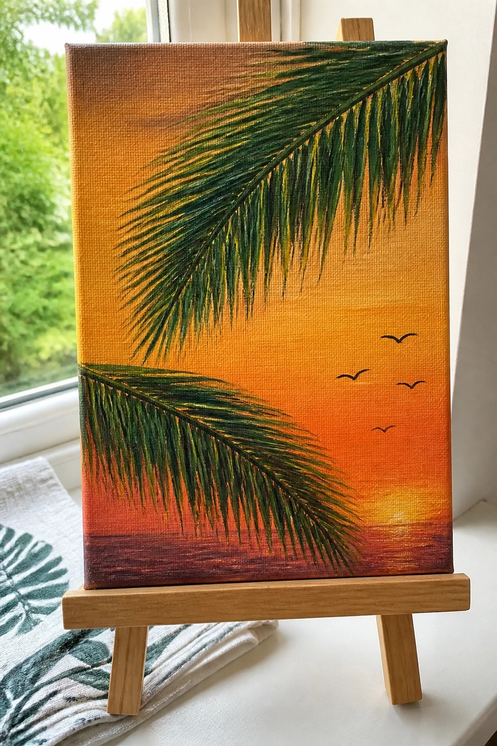 A small acrylic painting on a wooden easel shows two green palm fronds against an orange and red sunset sky with a reflective water line and several small black birds.
