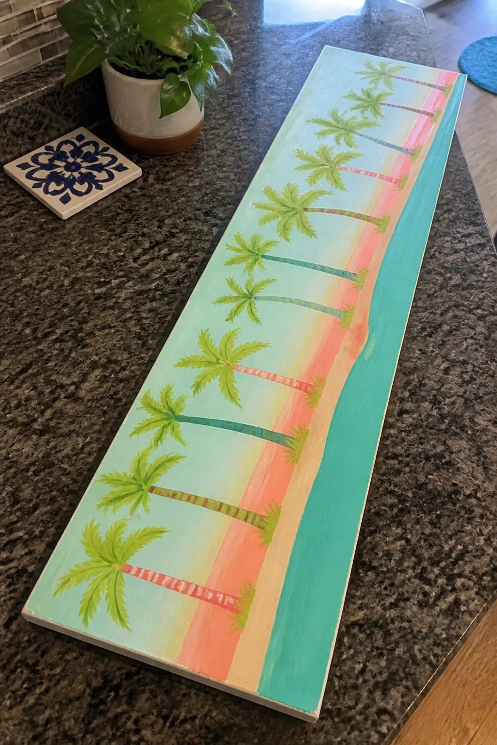 Colorful beach painting with palm trees on long wooden board beside plant