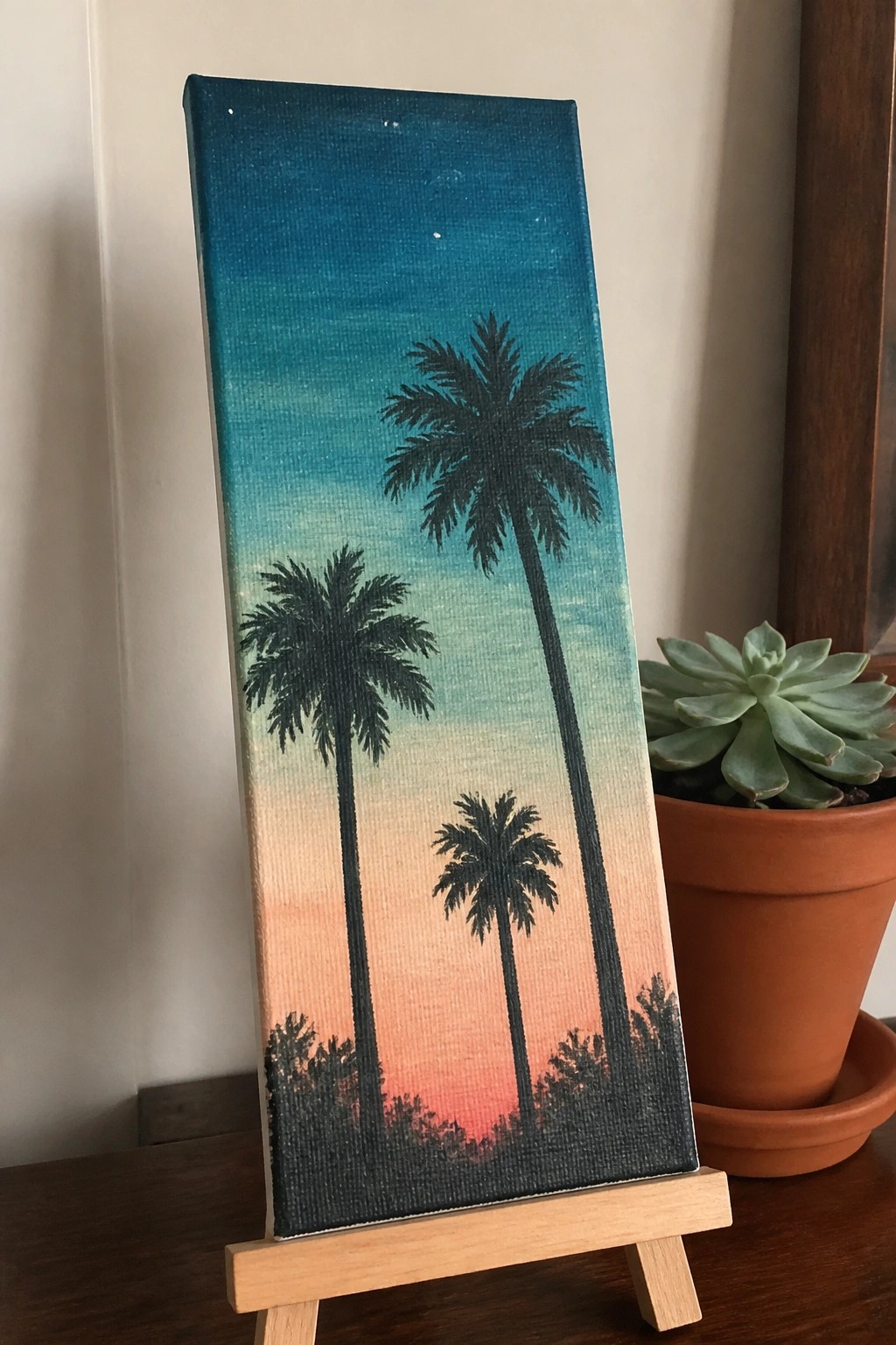Vertical canvas on easel with silhouetted palm trees against blue-to-orange sunset gradient beside succulent plant