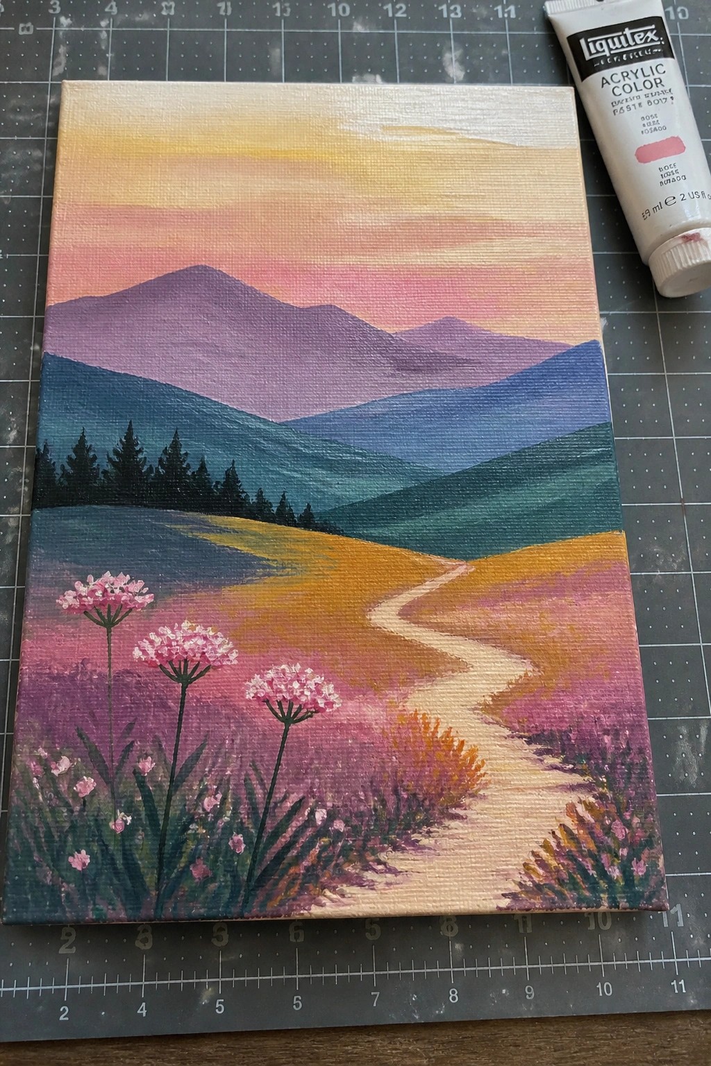 Vibrant acrylic mountain landscape painting with winding path and pink flowers beside paint tube