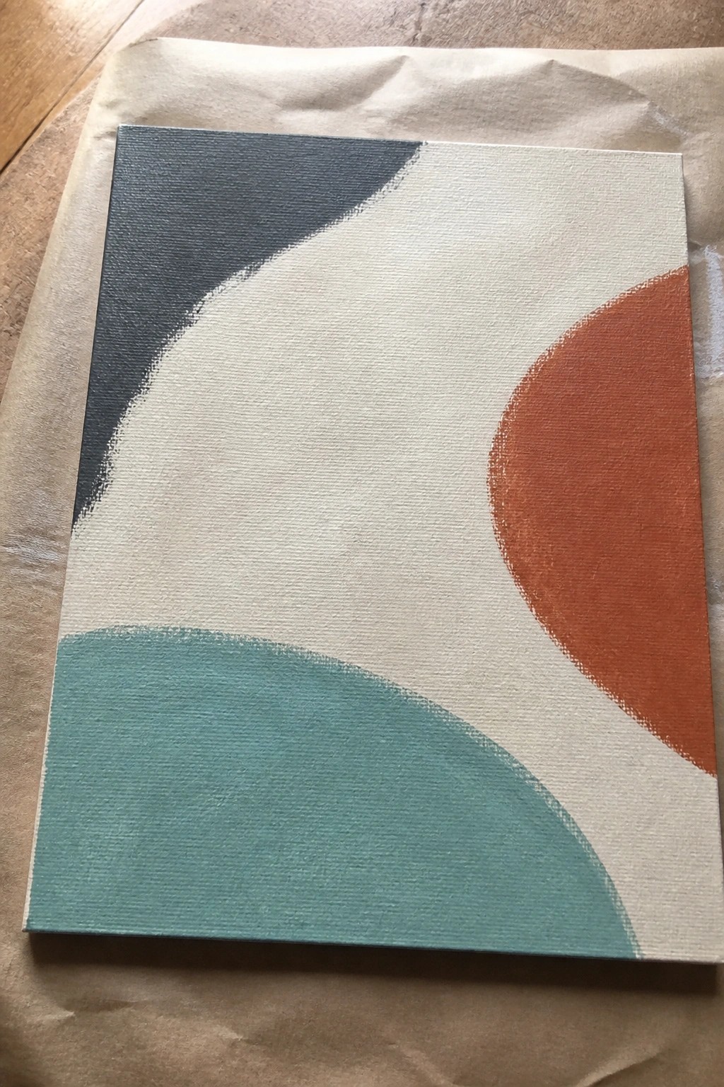 Abstract canvas with curved teal, blue, and orange shapes on white, atop brown paper.