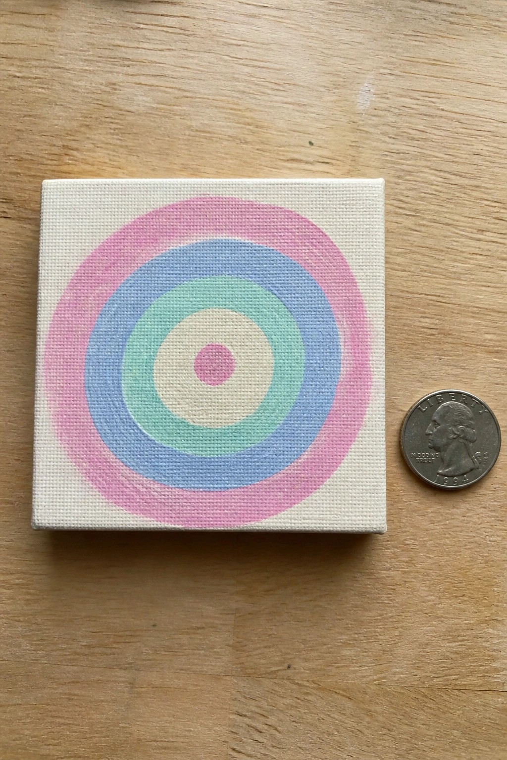 Small canvas with pastel pink blue green concentric rings beside a 1994 quarter.