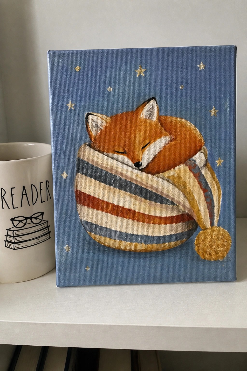 Sleeping fox curled in striped hat on starry blue canvas beside reader mug.