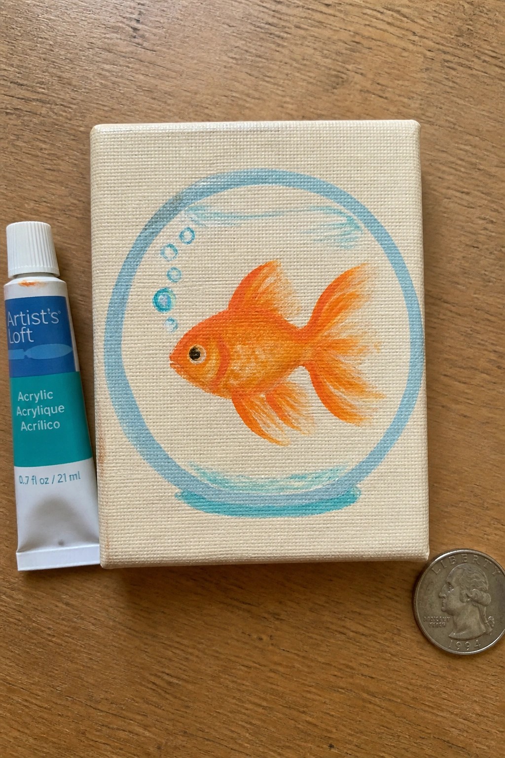 Orange goldfish painting on mini canvas beside acrylic tube and quarter on wood