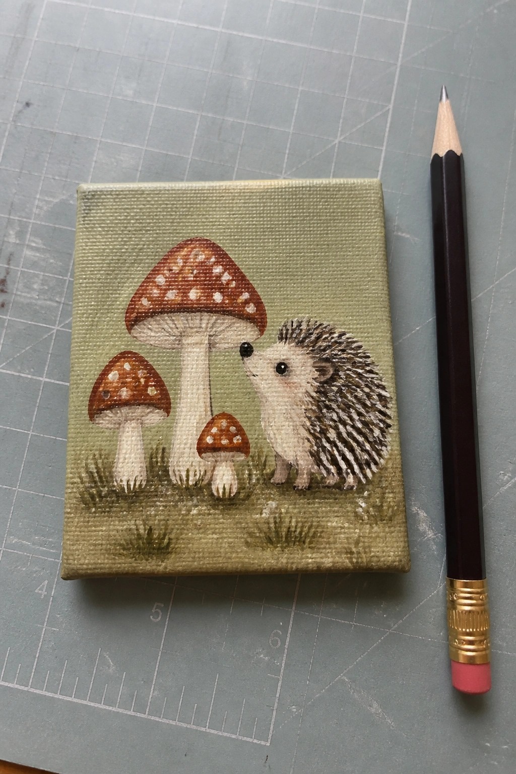 A small acrylic painting of a white hedgehog next to three brown spotted mushrooms on a green background with grass details, shown beside a pencil for scale.