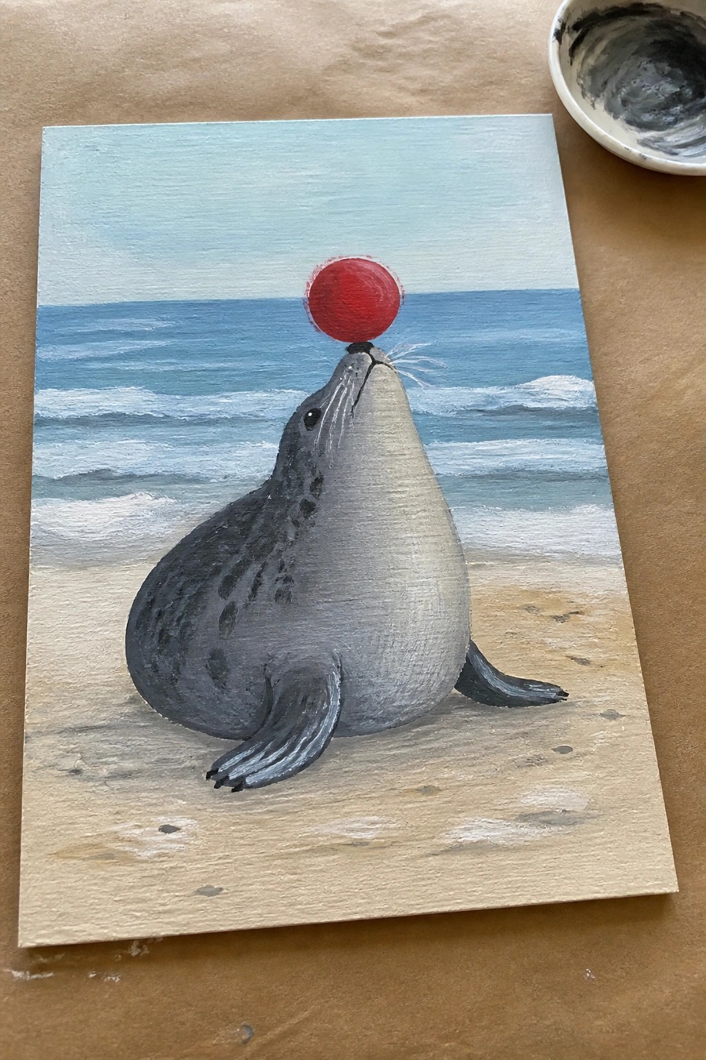 Gray seal balances red ball on nose on sandy beach with ocean waves.
