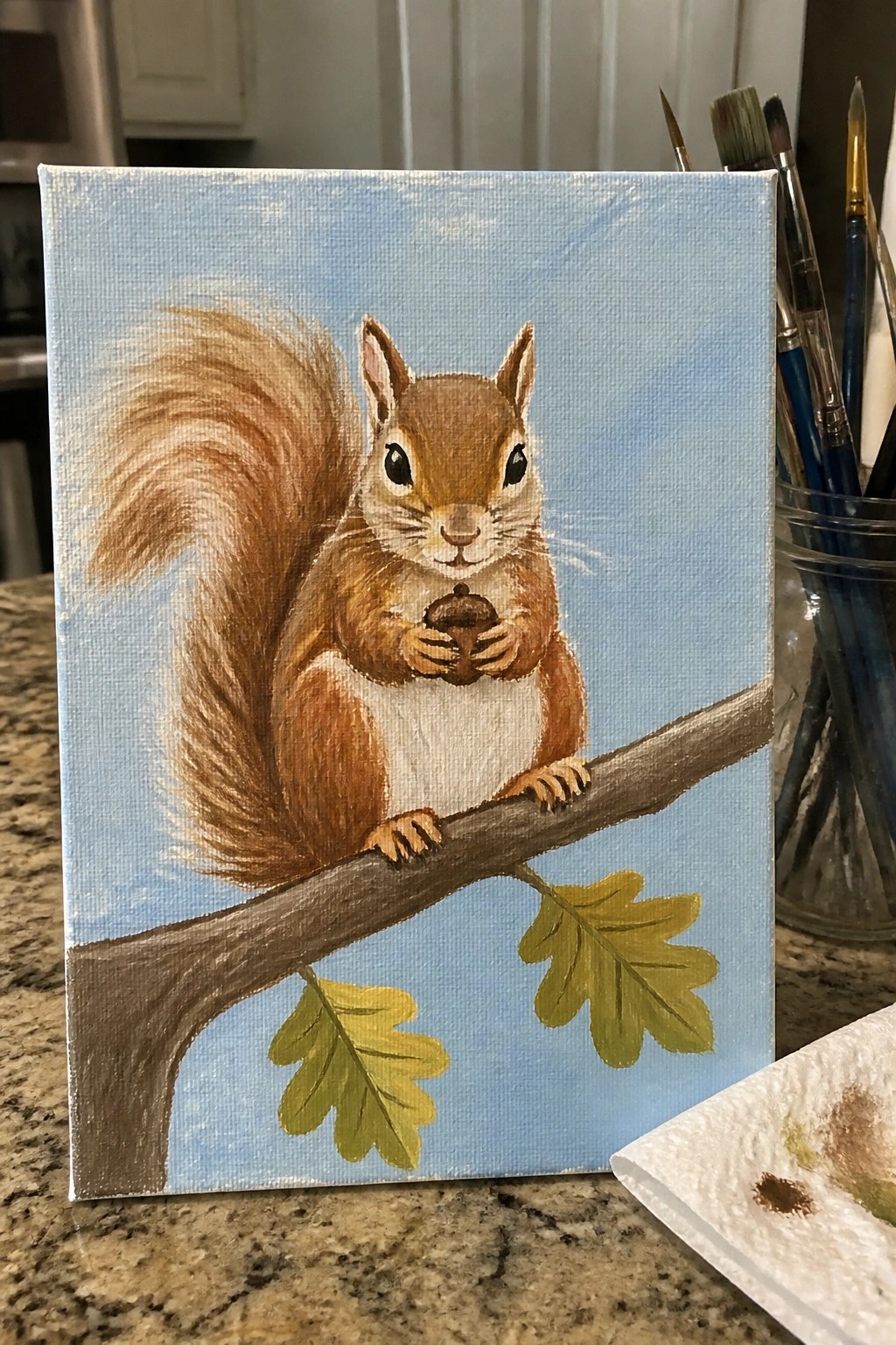 Brown squirrel holding acorn on branch with green leaves against blue sky.