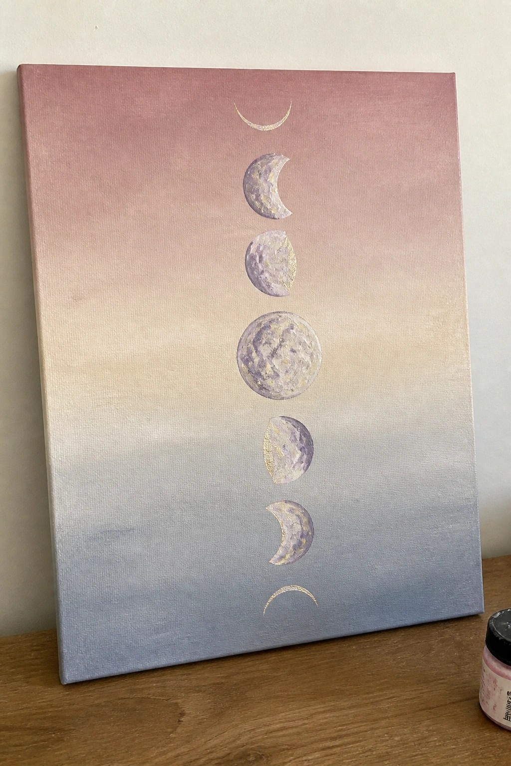 Canvas with moon phases on pink-blue gradient background on wooden table beside paint jar.