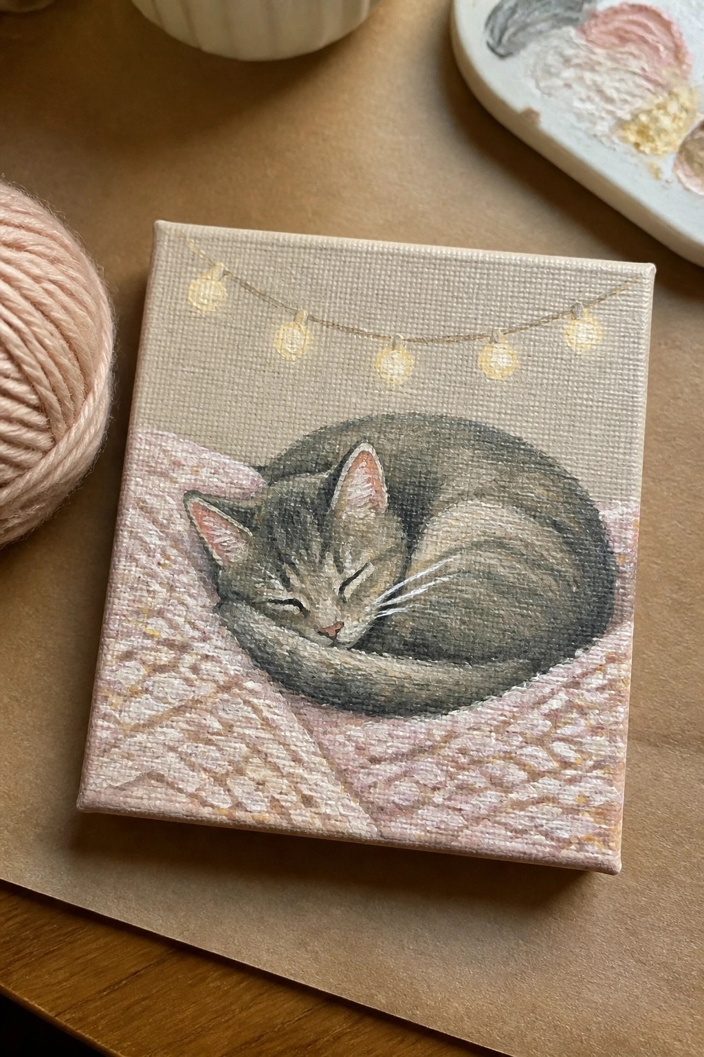 Miniature painting of sleeping gray cat curled on pink knit blanket under string lights