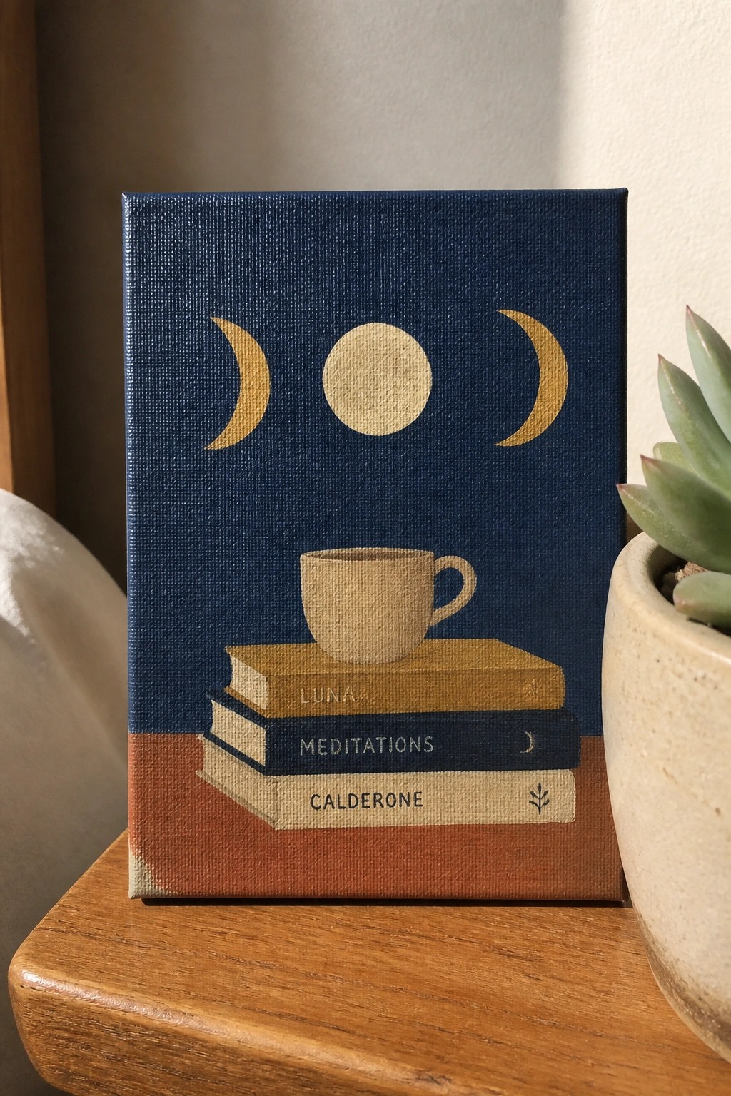 Blue canvas with gold moons above stacked books and coffee cup beside succulent