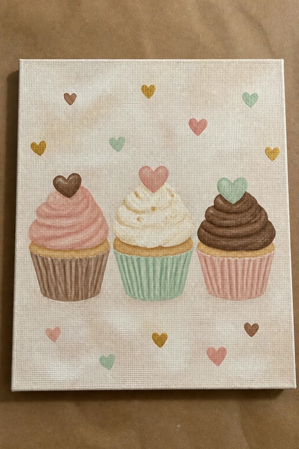 Three cupcakes with pink, white, and brown frosting topped with hearts amid scattered pastel hearts