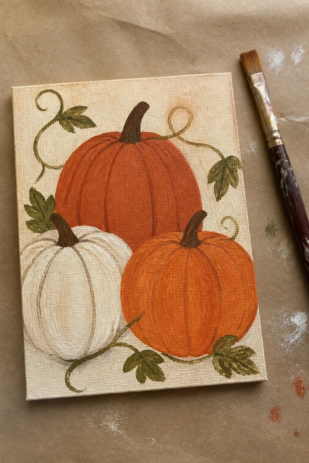 Acrylic painting of three pumpkins with green vines on canvas beside paintbrush