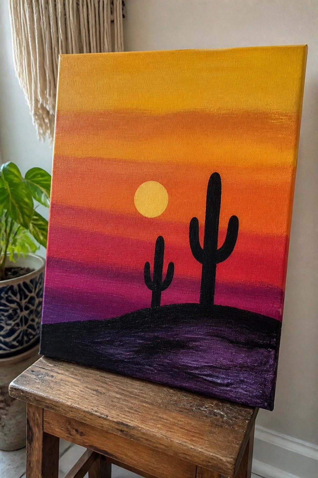 Sunset painting with yellow sun, two black cacti on purple hill atop wooden stool.