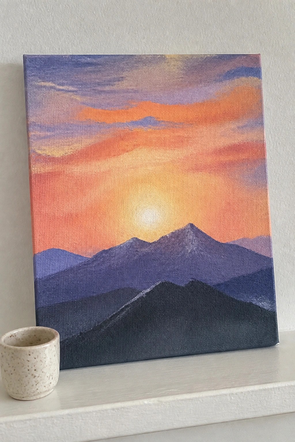 Vibrant sunset painting with layered purple mountains beside speckled ceramic cup on shelf