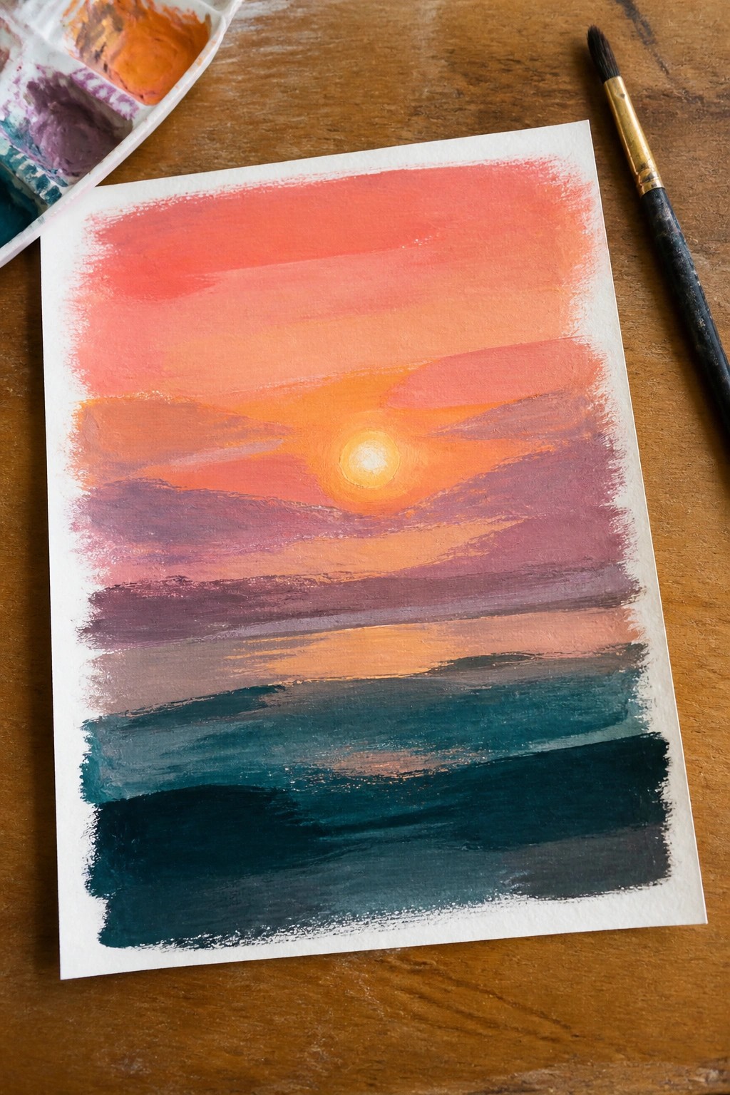 Watercolor sunset painting with glowing sun over layered orange purple teal beside palette and brush