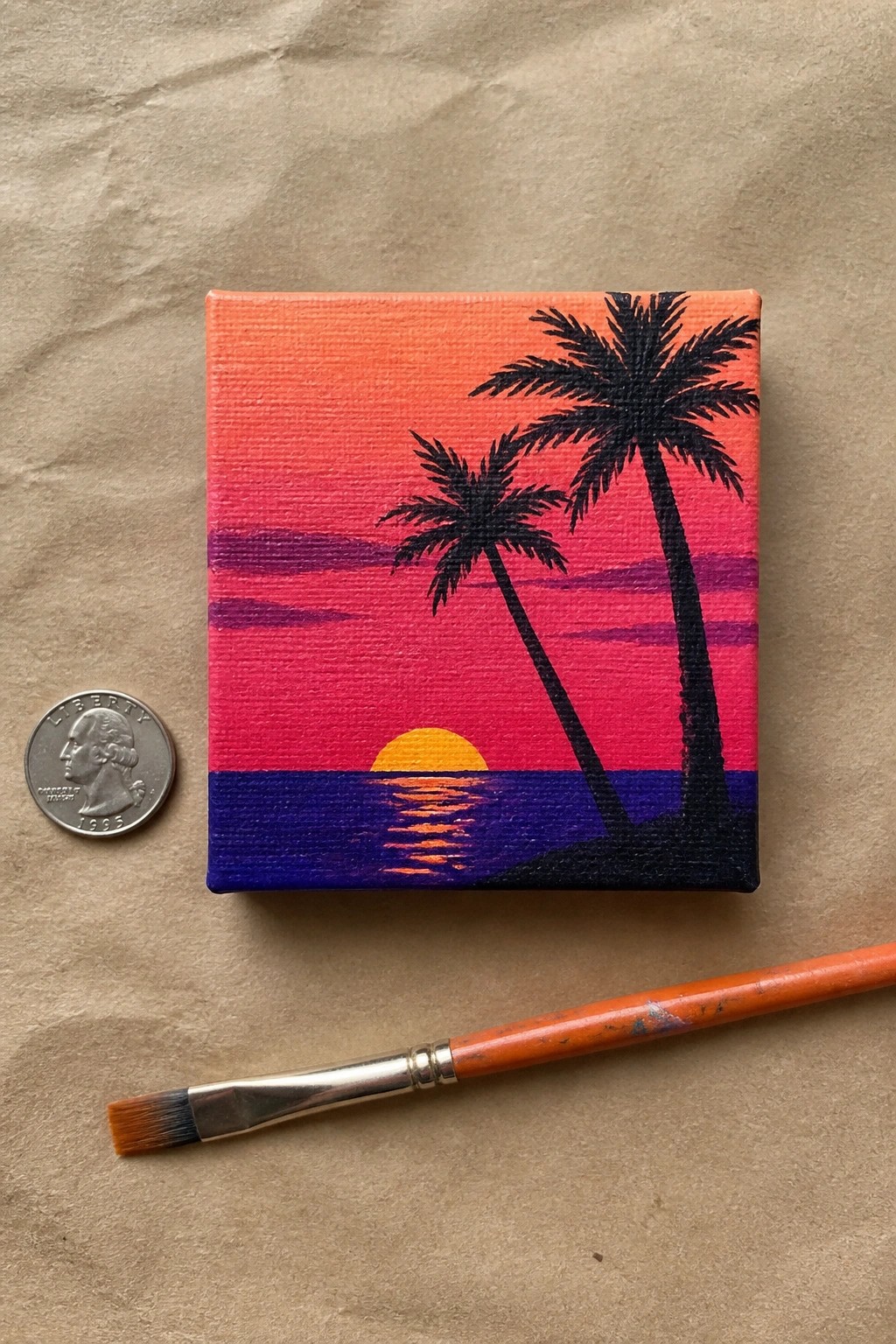 Tiny canvas sunset painting with palm trees beside quarter and orange brush on paper