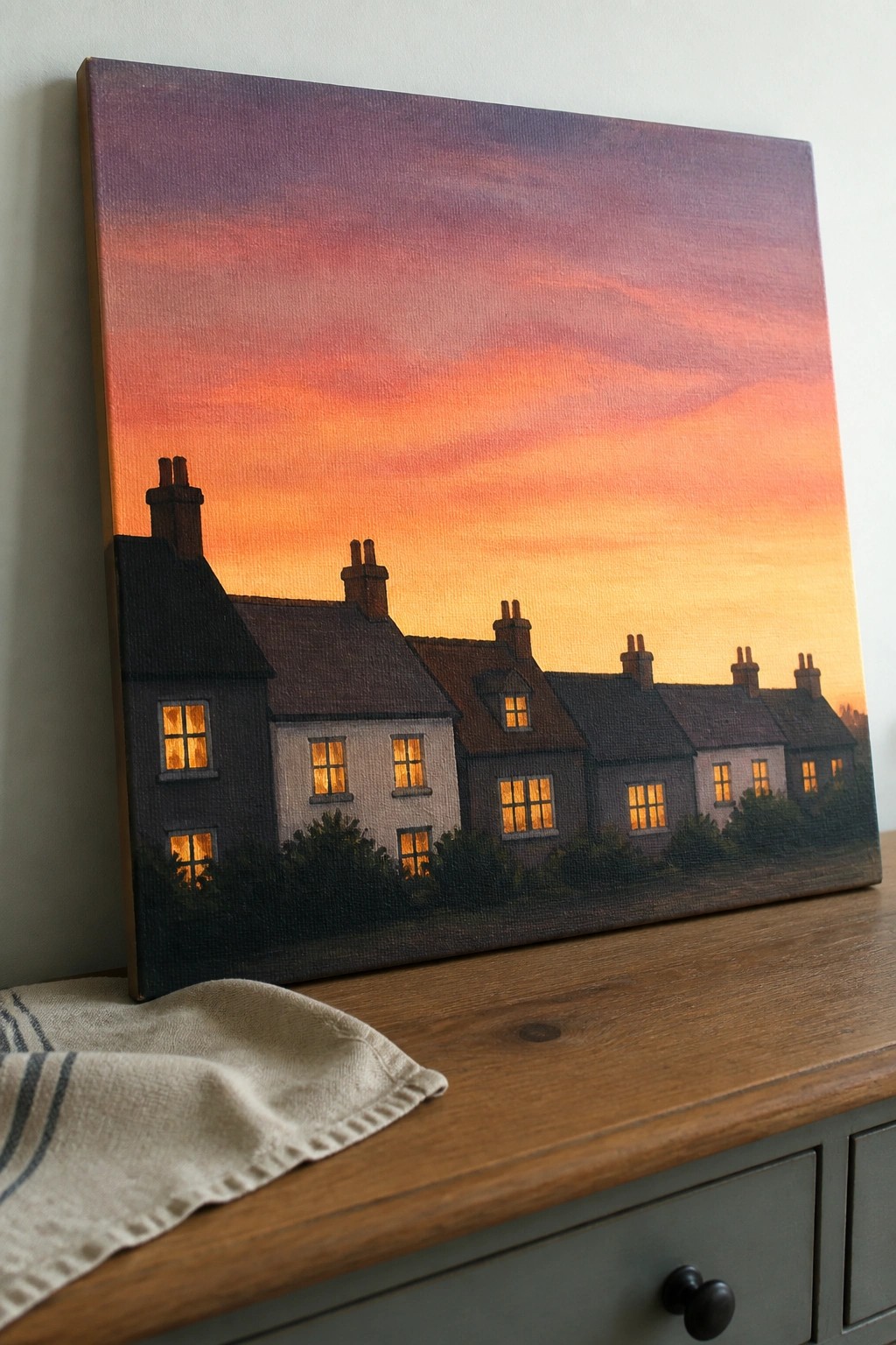 Painting of row houses with glowing windows under vibrant sunset sky on wooden dresser.