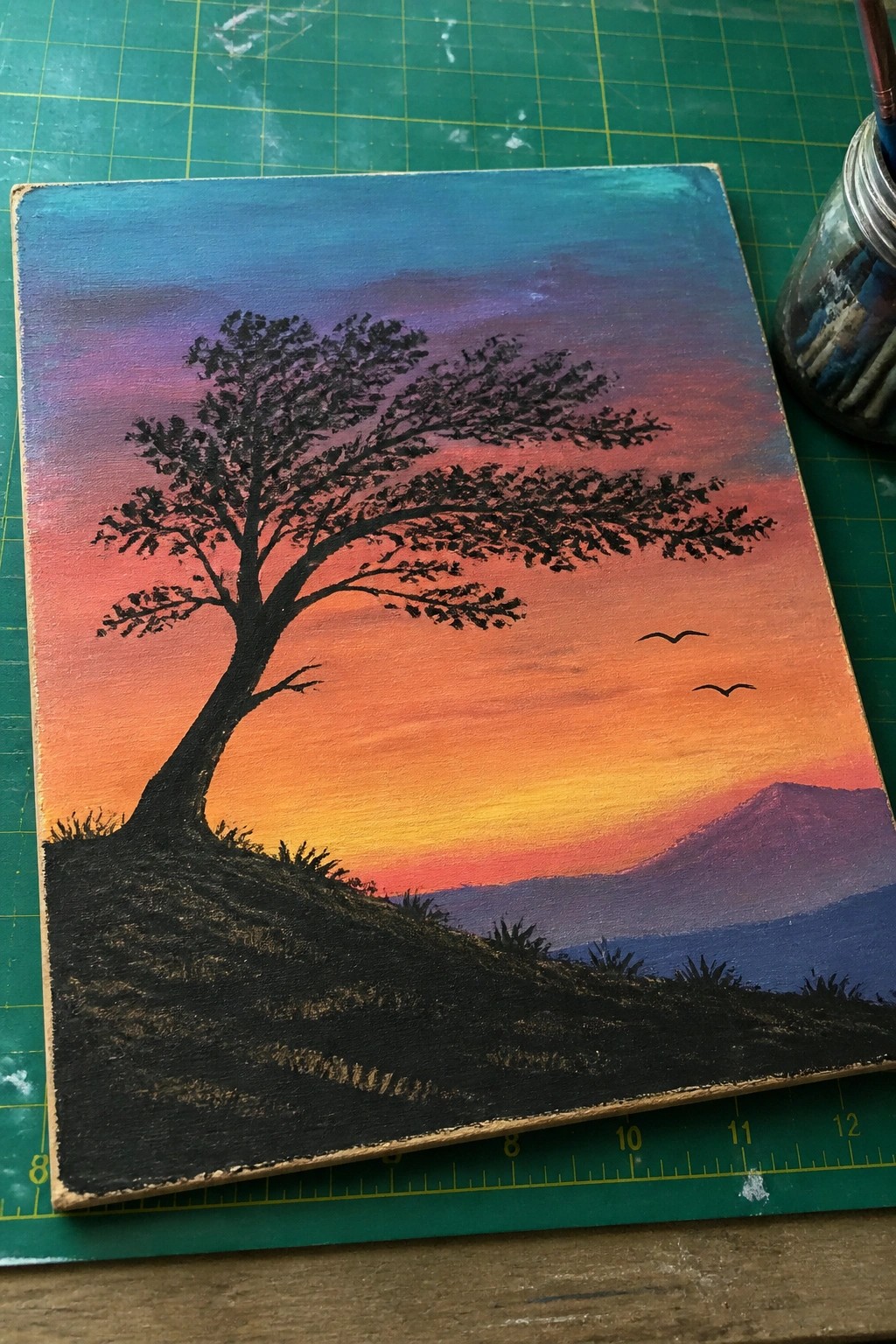 Silhouetted tree on hillside beneath vibrant orange-purple sunset sky with birds and mountains