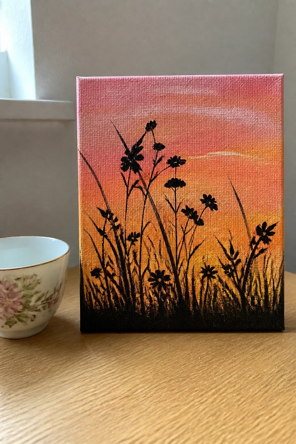 Black flower silhouettes on pink-orange sunset canvas beside floral teacup on wood table.
