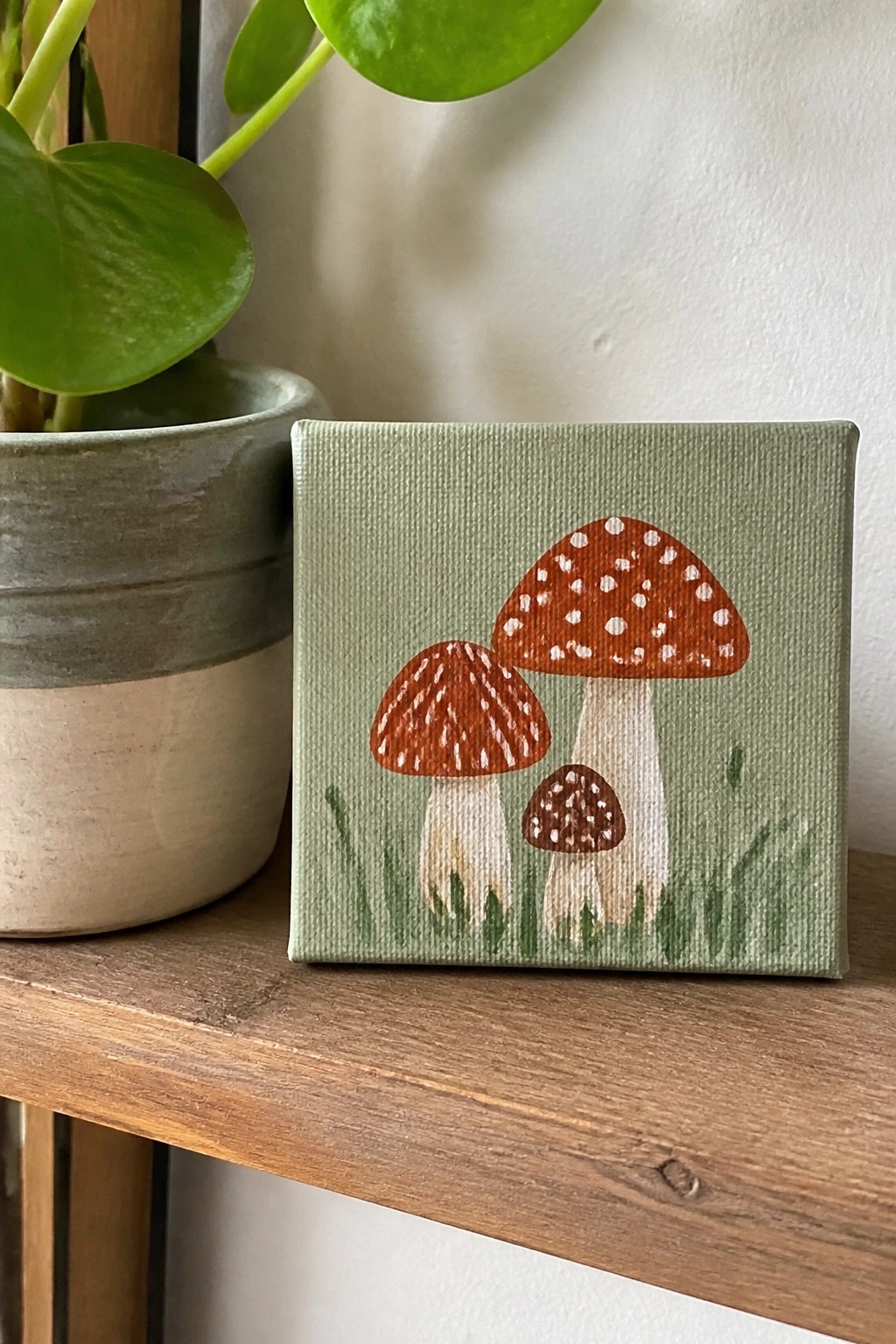 Small canvas with three red-spotted mushrooms on light green background beside plant pot