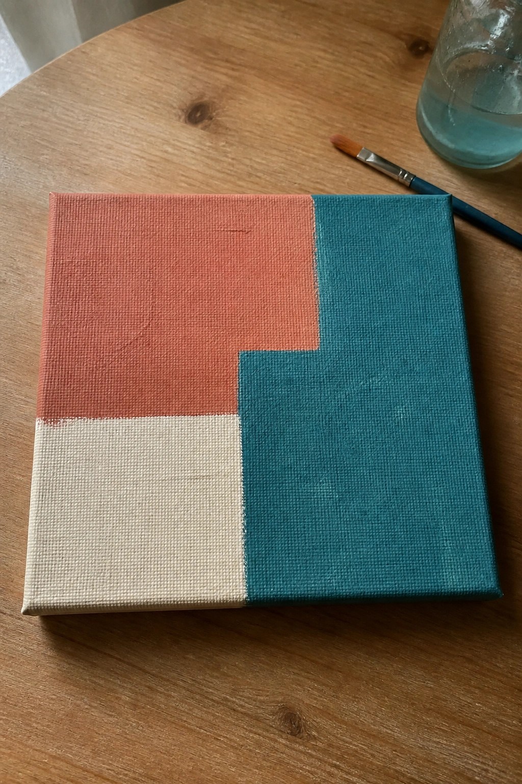 Geometric abstract painting in coral, cream, and teal on wooden table with brush.