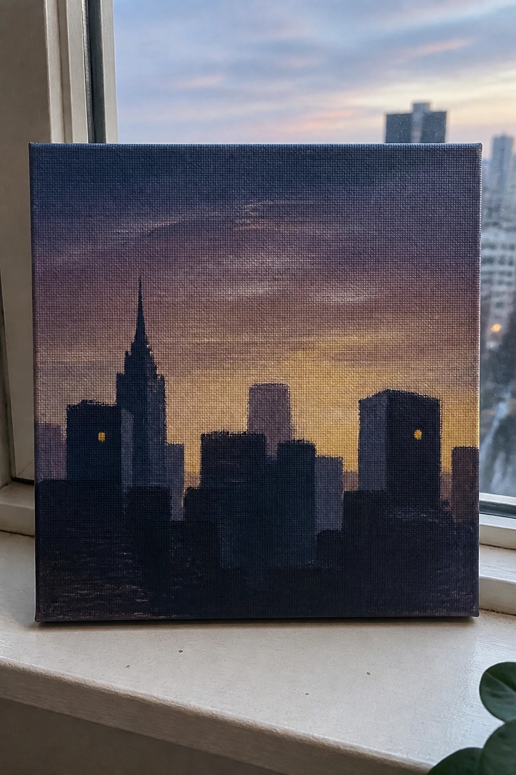 Painting of dark skyscrapers silhouetted against purple-orange sunset sky on windowsill