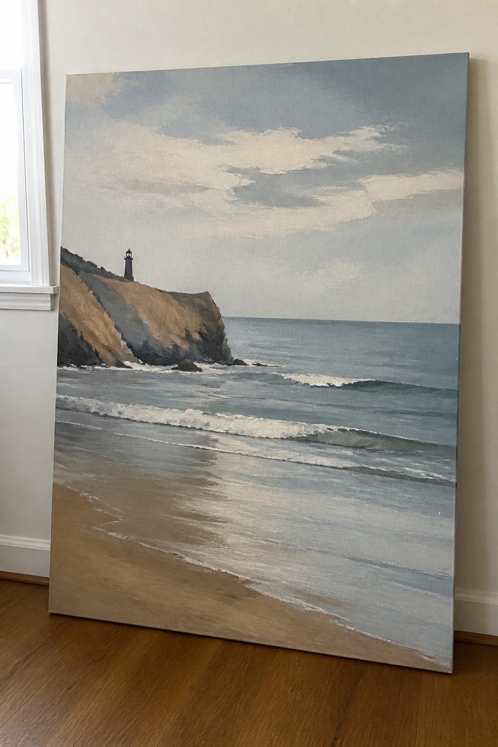 Large seascape painting of lighthouse on cliff above wavy ocean and sandy beach indoors