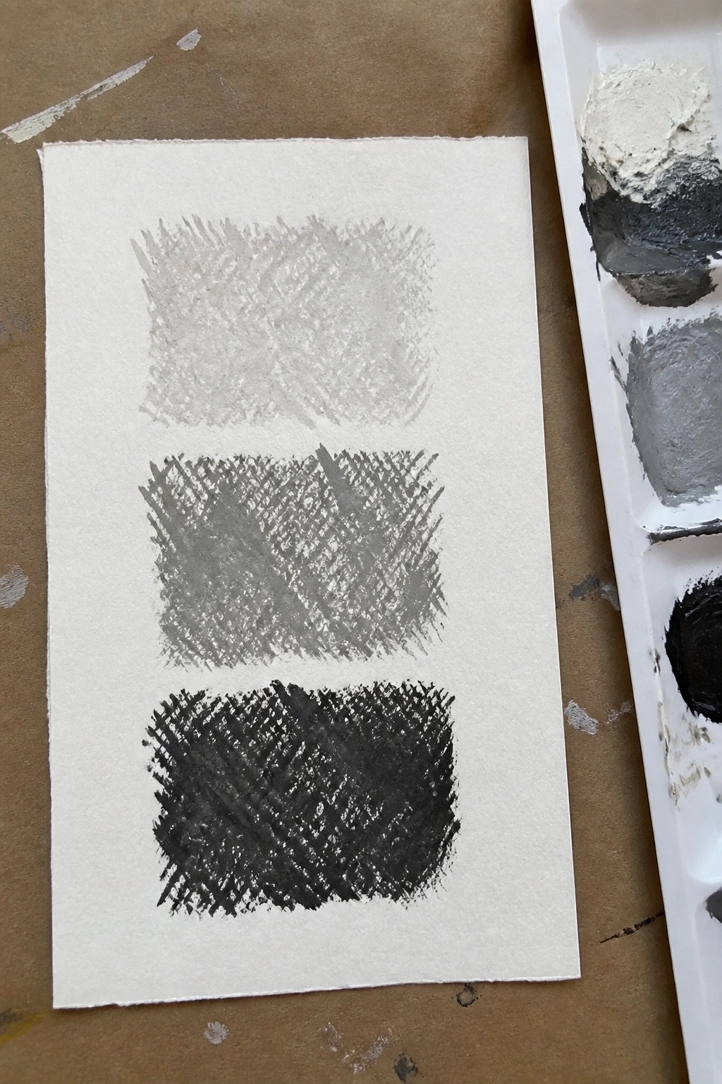 Three graduated gray swatches on white paper beside a paint palette.