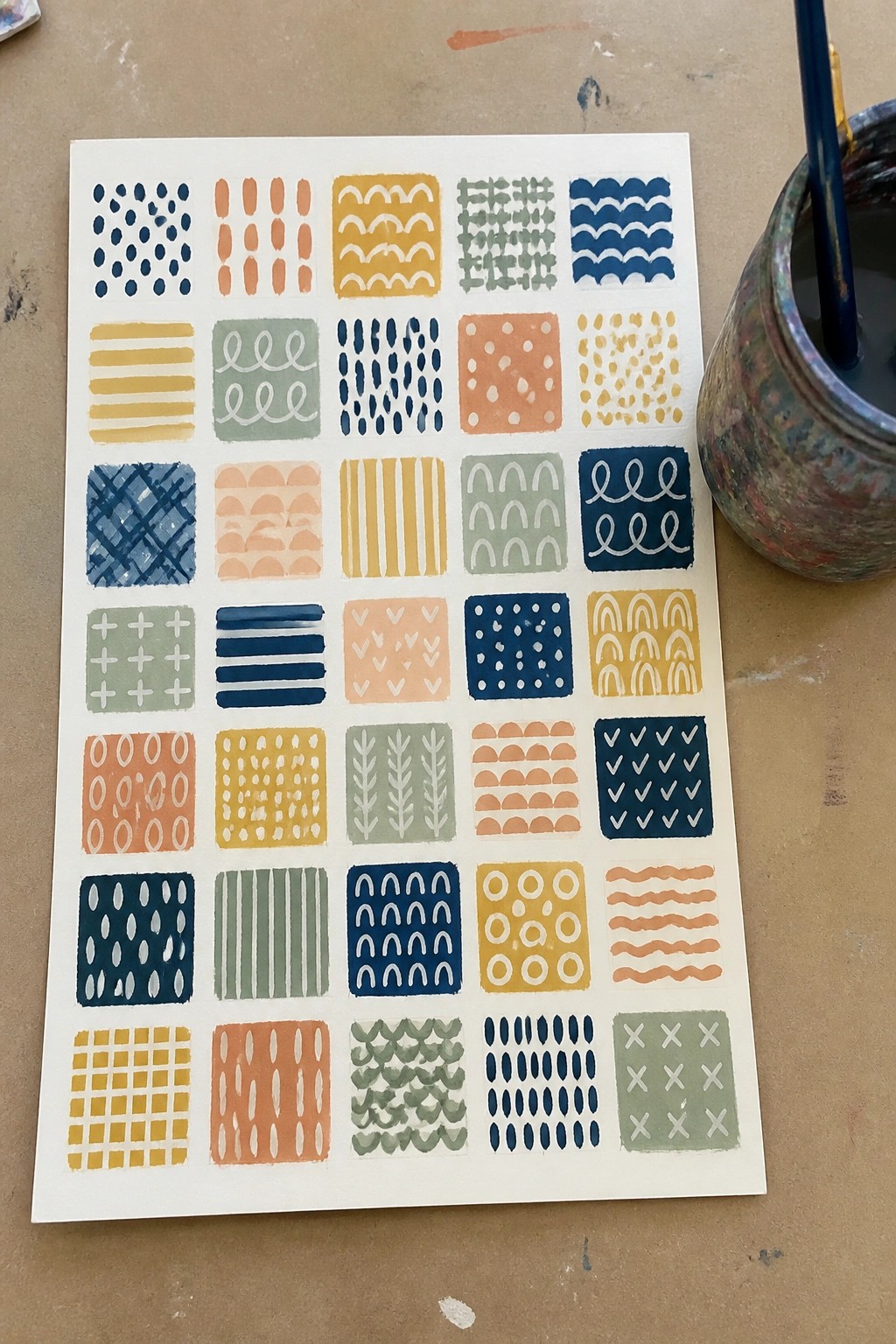 Grid of colorful hand-painted geometric patterns on paper beside paint jar and brush