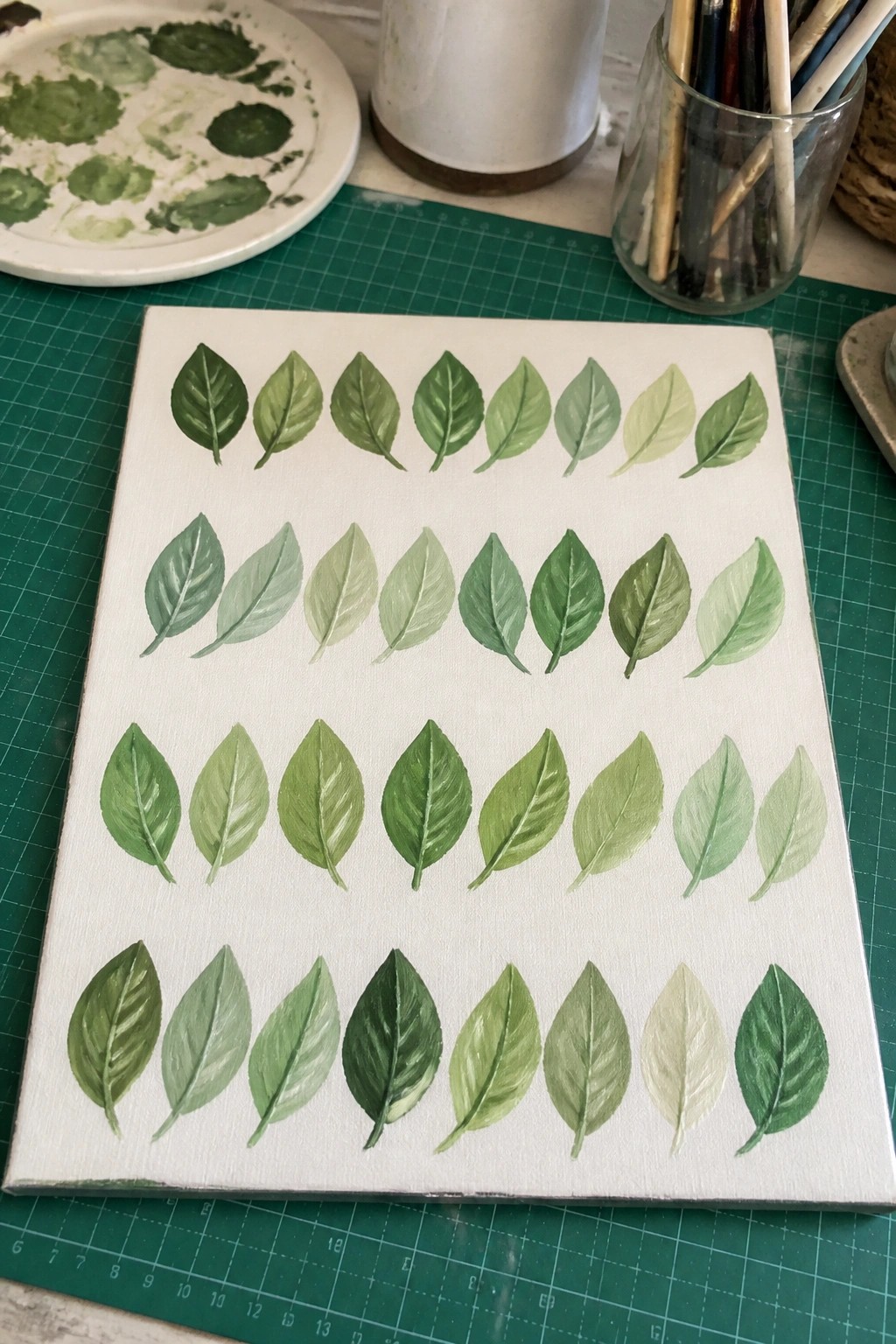 A white canvas on a green mat shows four horizontal rows of painted leaves in multiple shades of green, surrounded by a paint palette and brush jar.