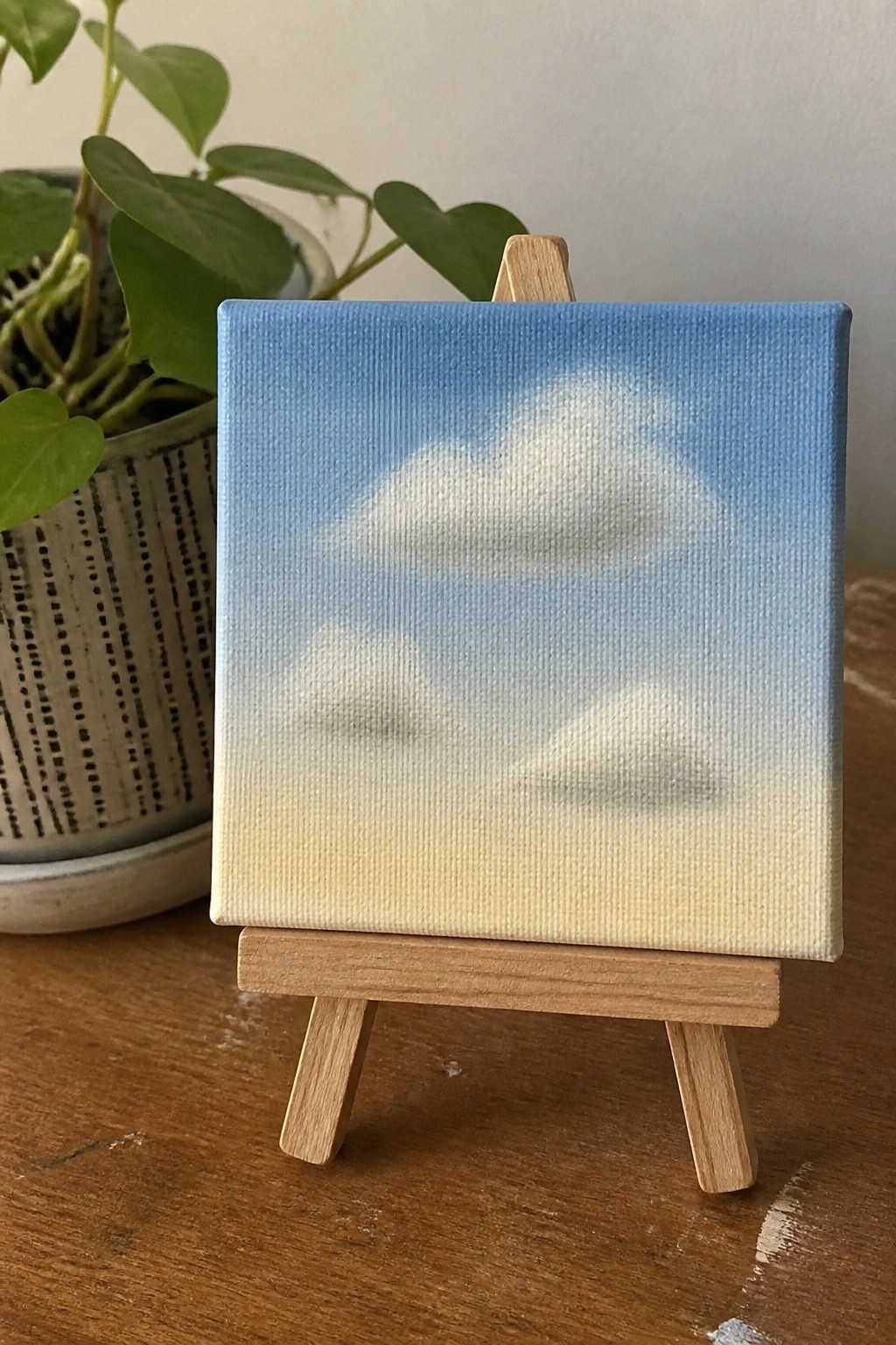 Small canvas on wooden easel shows blue sky with white clouds over yellow base