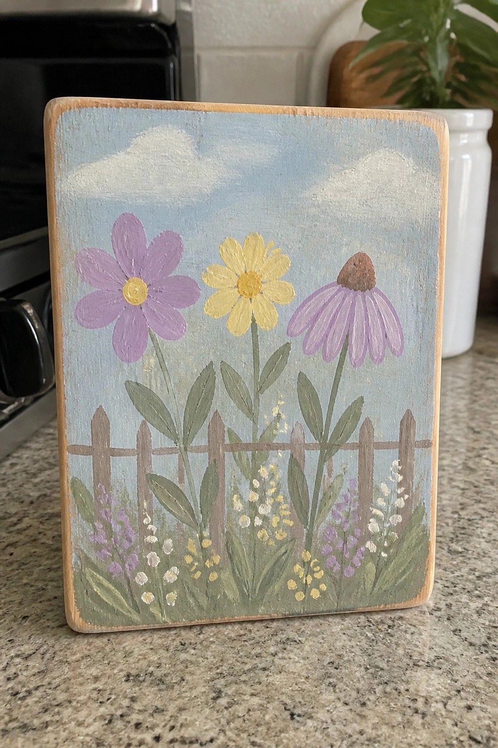 Wooden plaque painted with purple and yellow flowers behind picket fence under blue sky