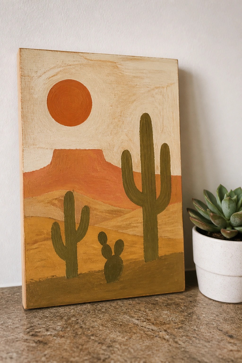Desert landscape painting with orange sun, red mountains, green cacti, and potted succulent.