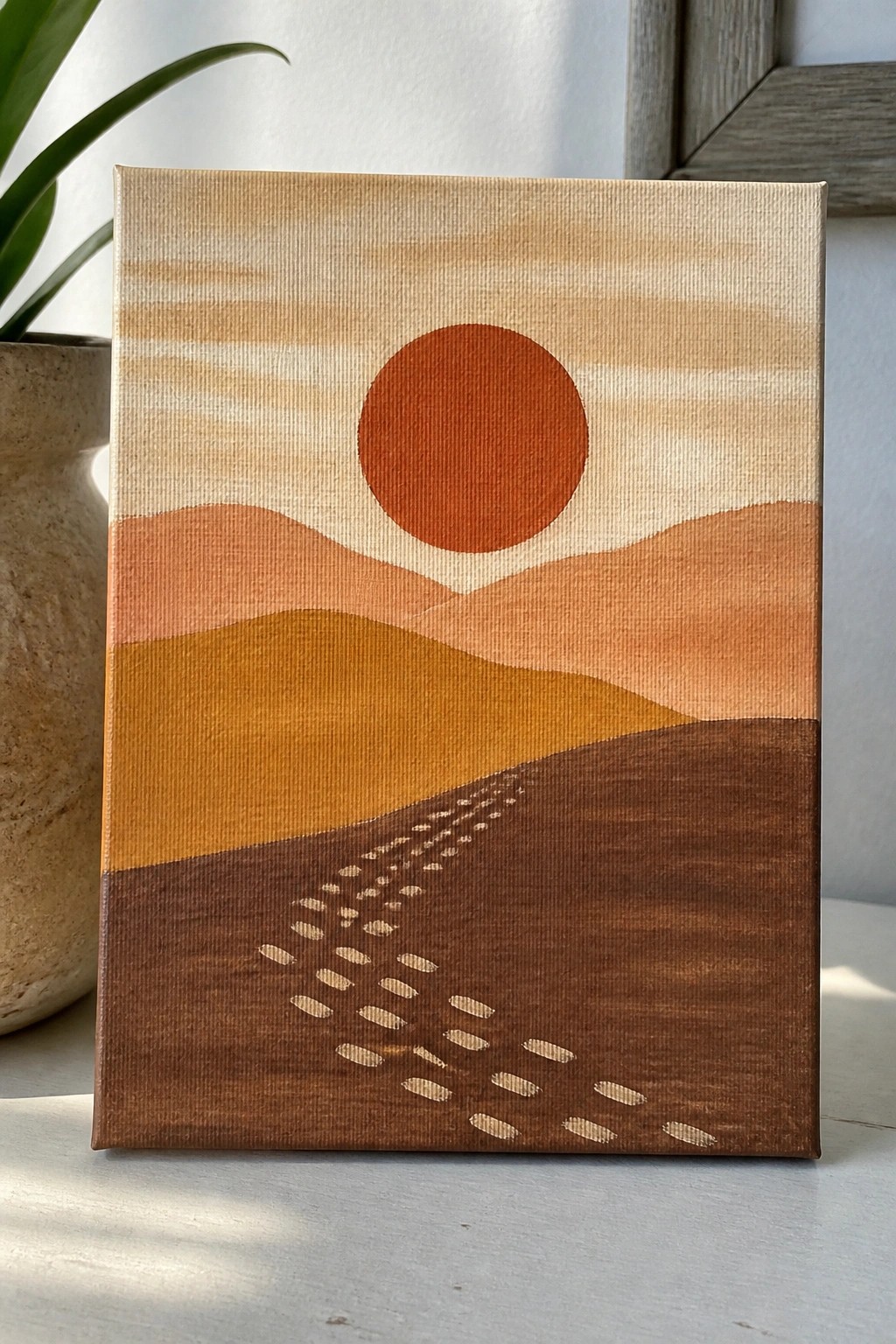 Minimalist canvas painting of orange sun over layered hills with footprints trail