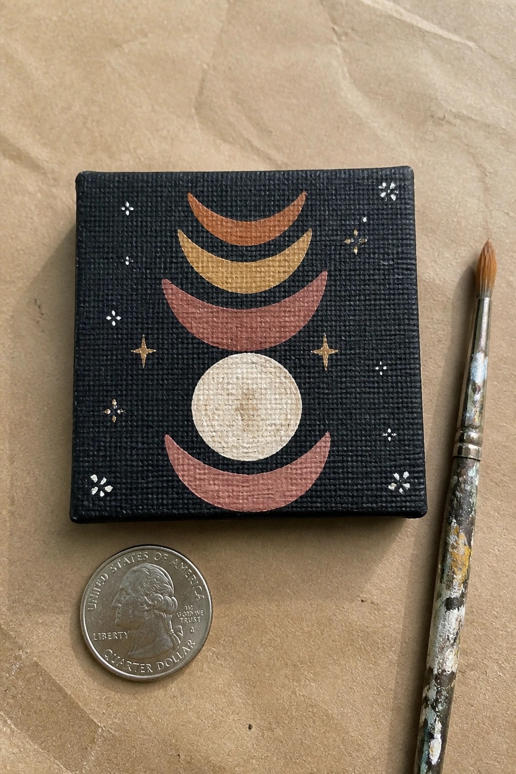 Miniature canvas with moon phases in earth tones beside quarter and paintbrush.