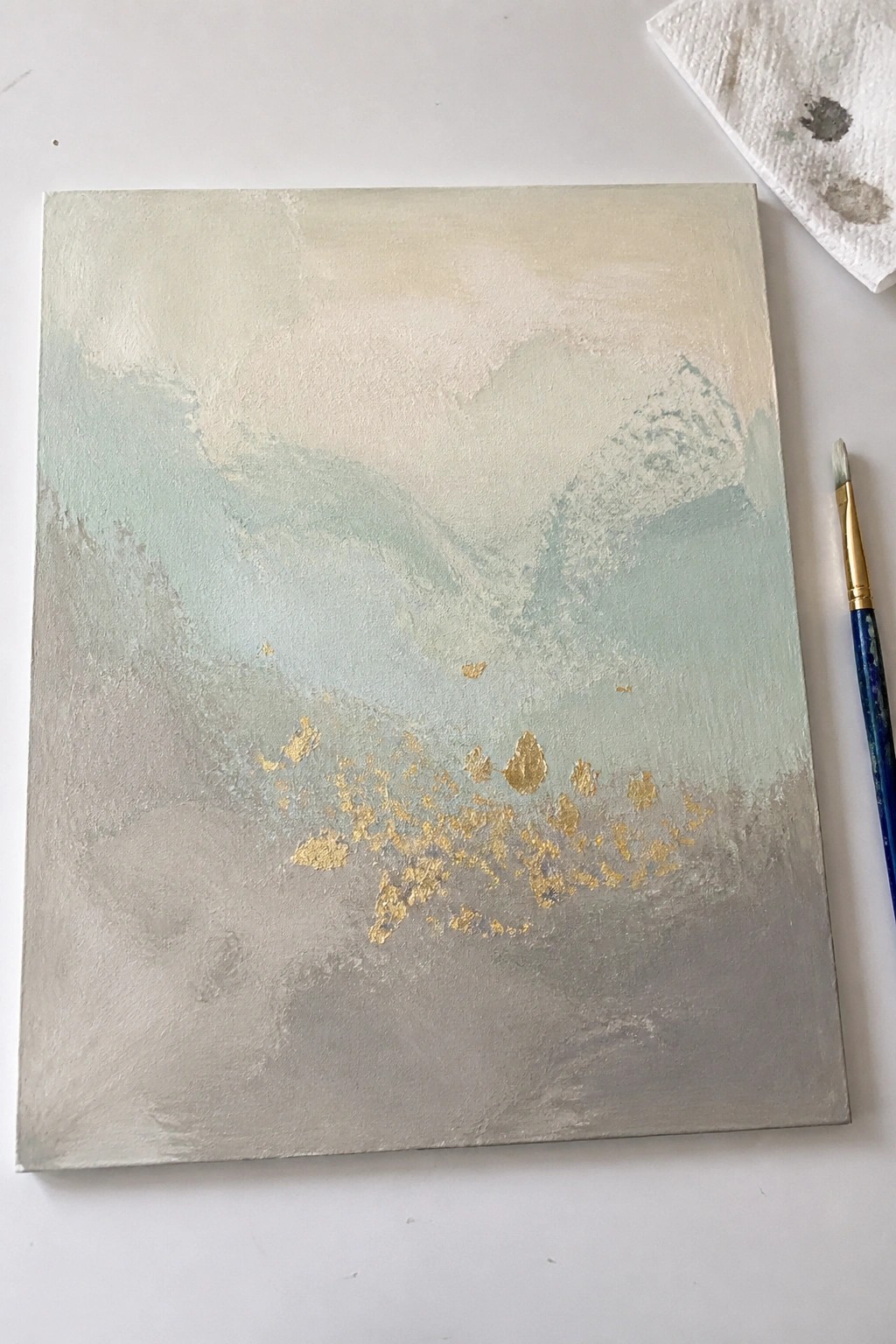 Abstract painting with soft green-gray tones, gold leaf accents, and nearby paintbrush.