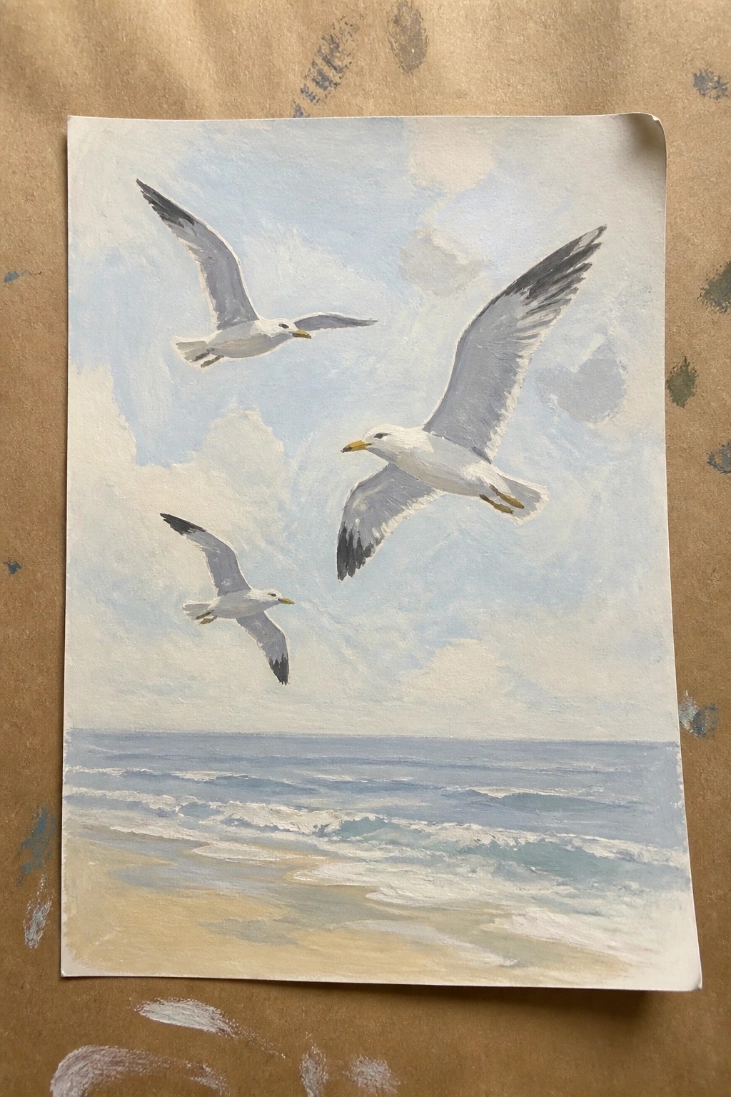 Three seagulls soar above blue ocean waves crashing onto a sandy beach.