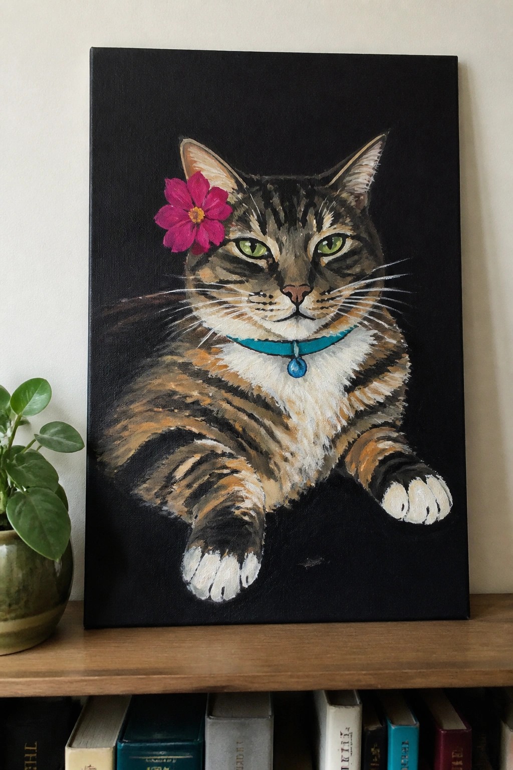 Tabby cat painting with pink flower, blue bell collar on black canvas above books