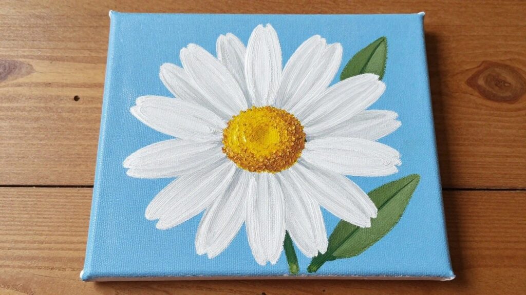 Small square canvas with white daisy, yellow center, and green leaves on blue background on wood.