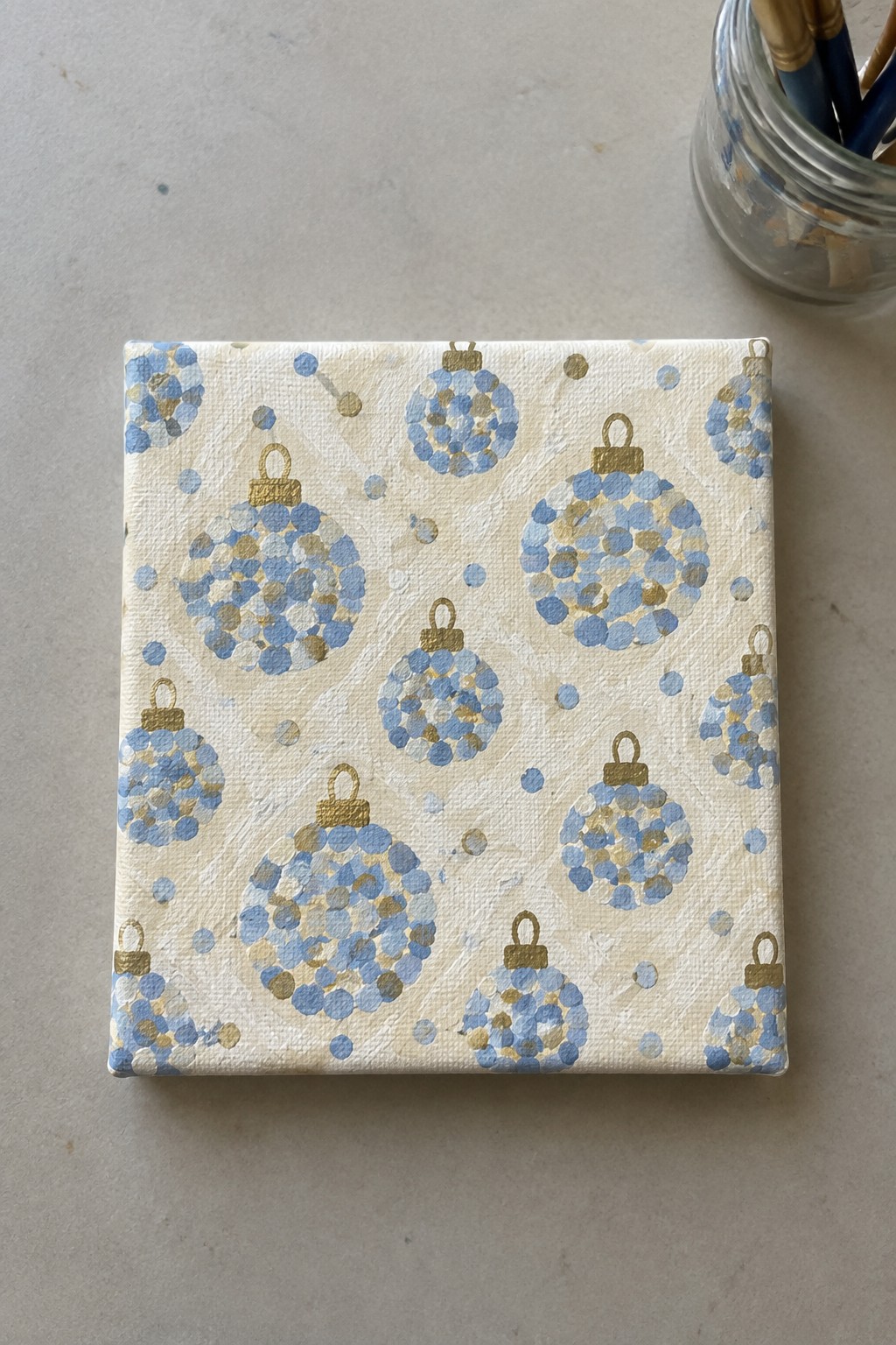 Square canvas with blue and gold dotted Christmas ornaments on beige background, brushes nearby.
