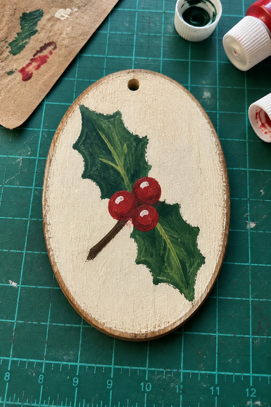 Oval wooden ornament painted with green holly leaves and red berries on cutting mat